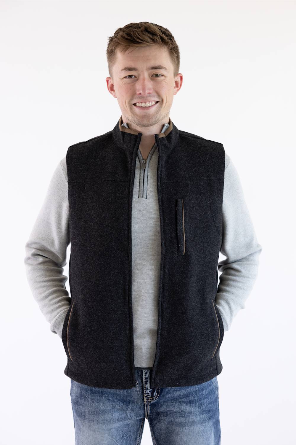 A smiling man wearing a dark charcoal fleece vest over a light gray quarter-zip sweater and blue jeans, standing against a plain white studio background.