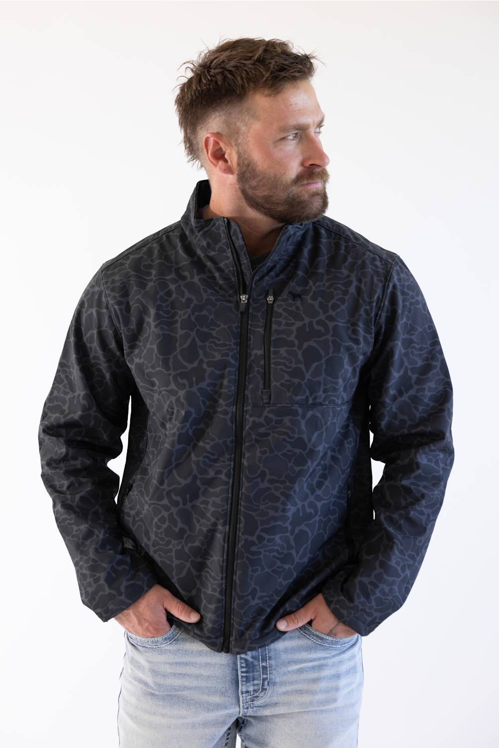 A man wearing a dark navy blue zip-up jacket with a subtle tonal abstract pattern, paired with light blue jeans, standing against a plain white background.