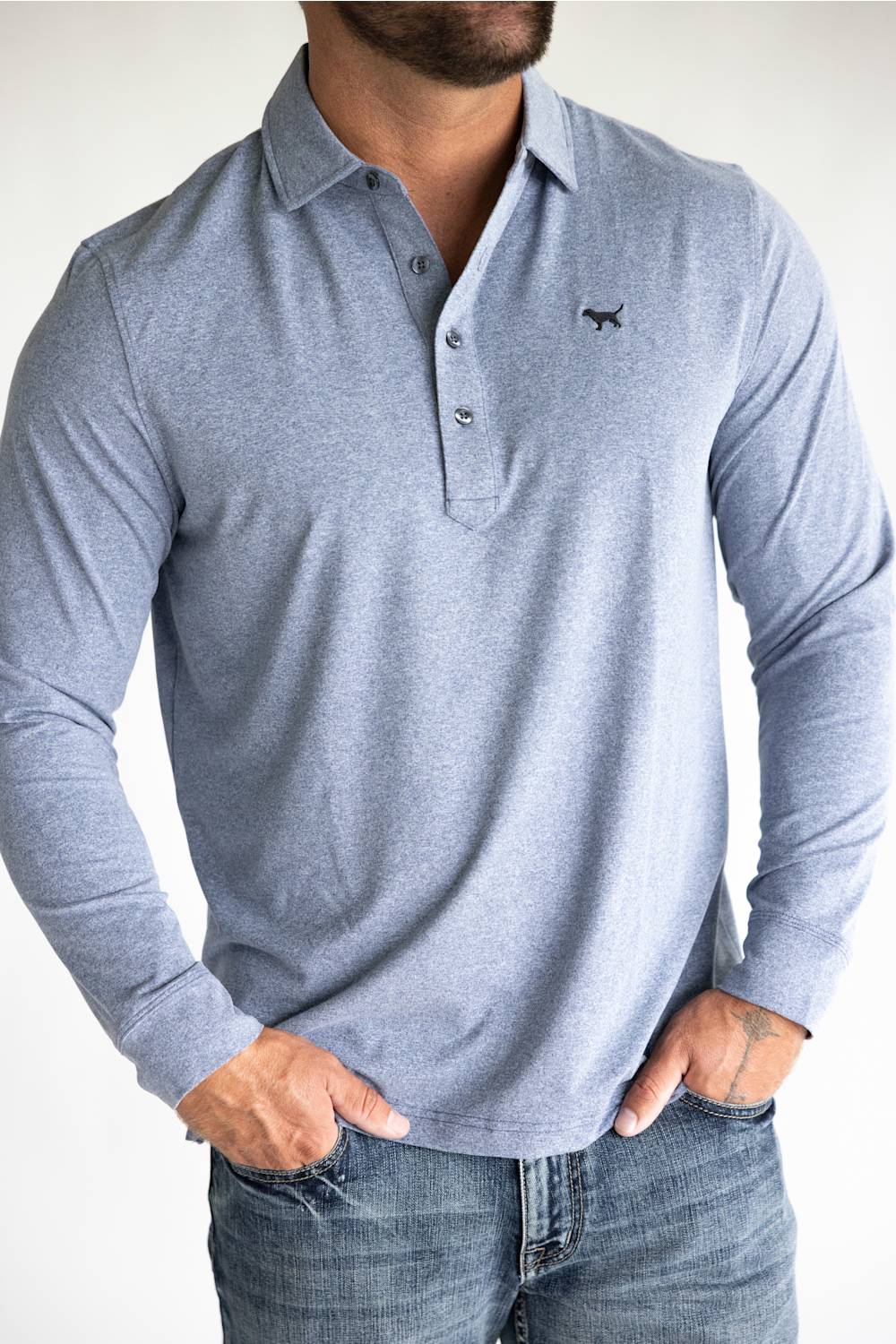 A man wearing a light heather gray long-sleeve polo shirt with a three-button placket and a small black embroidered dog logo on the left chest, paired with blue jeans.