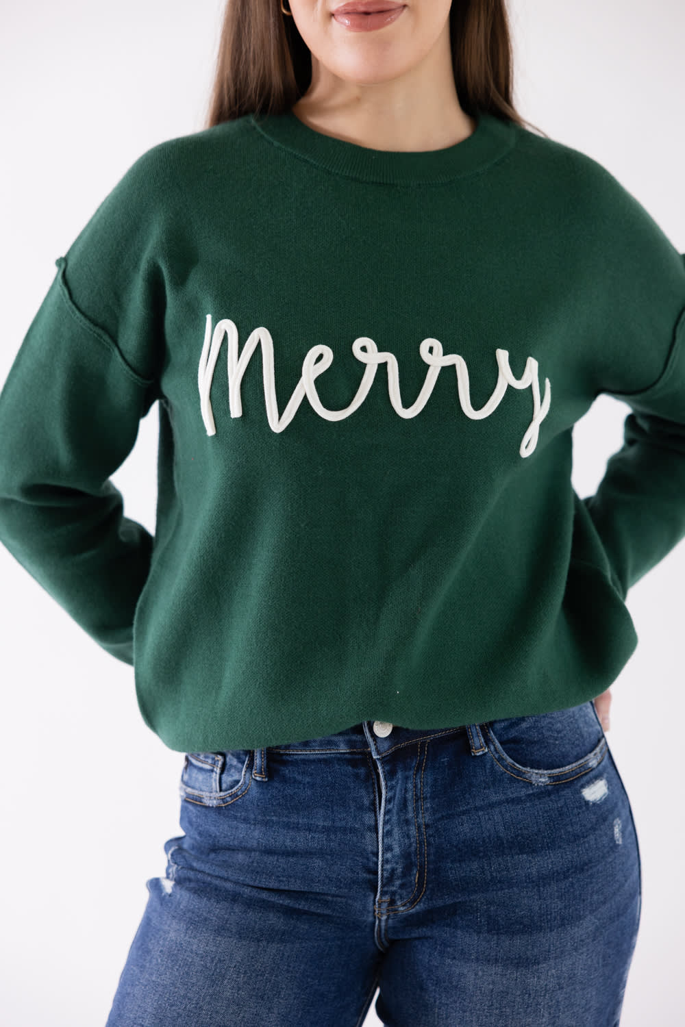 A woman wearing a dark green crewneck sweatshirt with 'Merry' written in white cursive script across the chest, paired with blue jeans.
