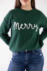 A woman wearing a dark green crewneck sweatshirt with 'Merry' written in white cursive script across the chest, paired with blue jeans.