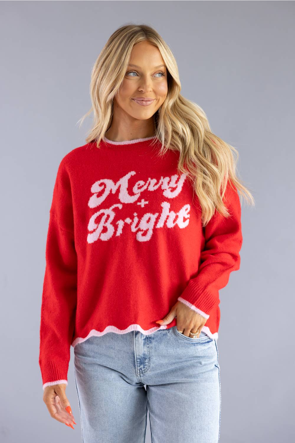 A woman wearing a bright red knit sweater with 'Merry Bright' written in white across the chest, paired with light blue jeans, against a solid gray studio background.