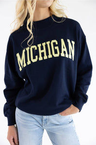 A woman wearing a navy blue crewneck sweatshirt with 'MICHIGAN' printed across the chest in a cream-colored varsity font, paired with light-wash blue jeans.