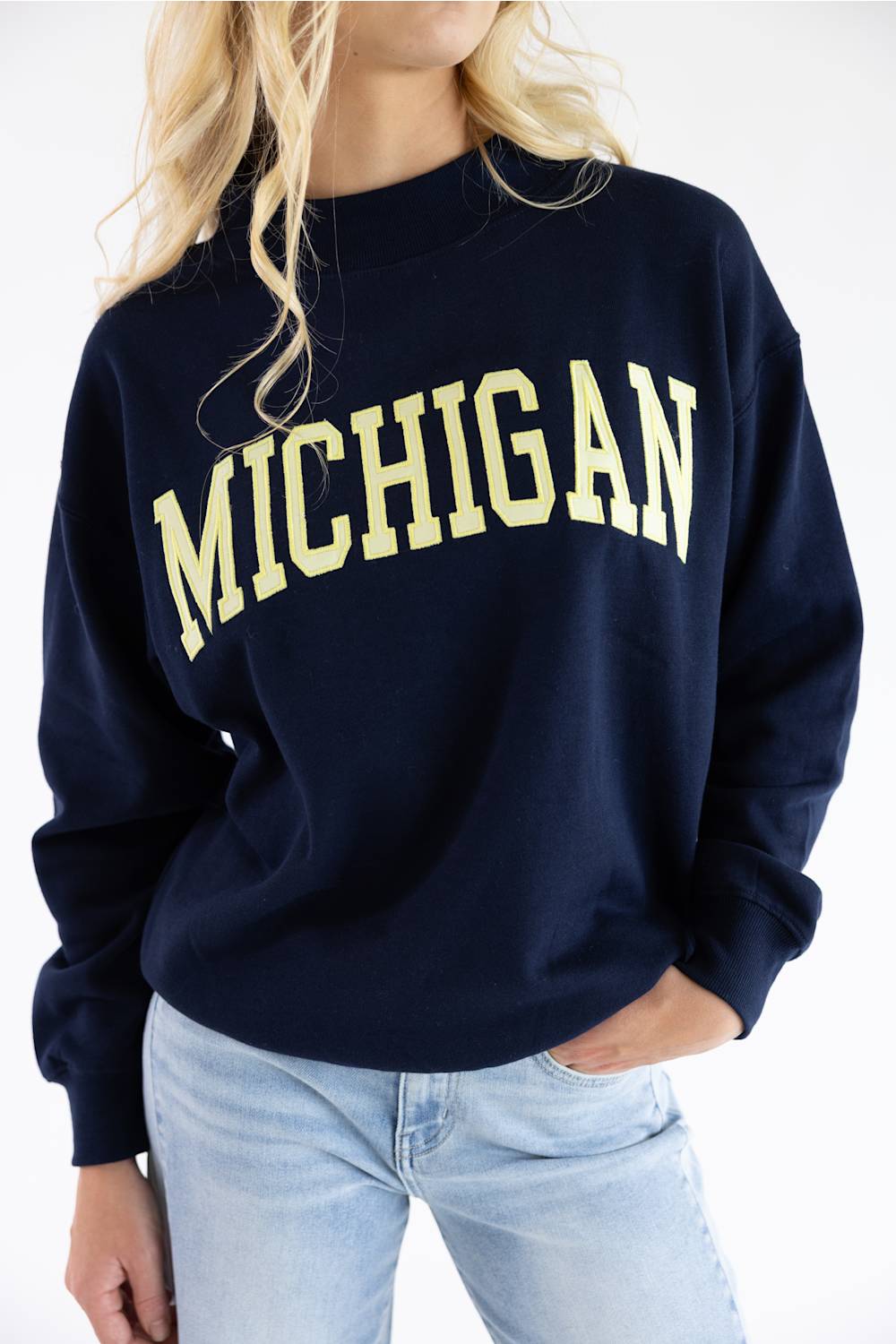 A woman wearing a navy blue crewneck sweatshirt with 'MICHIGAN' printed across the chest in a cream-colored varsity font, paired with light-wash blue jeans.