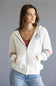 A woman wearing a white zip-up hoodie with colorful striped cuffs, a brown ribbed top, and blue jeans, photographed against a solid gray studio background.