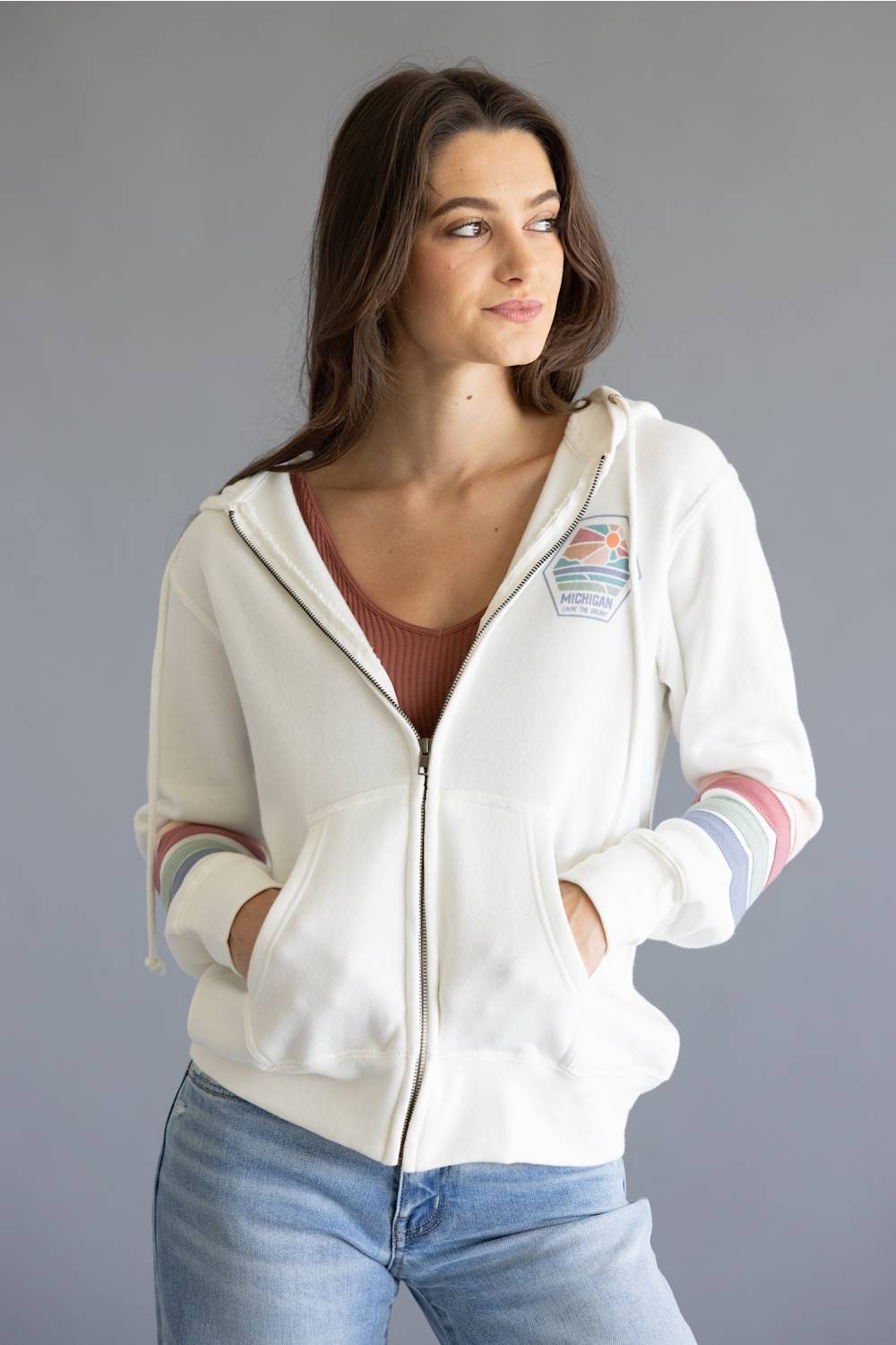 A woman wearing a white zip-up hoodie with colorful striped cuffs, a brown ribbed top, and blue jeans, photographed against a solid gray studio background.