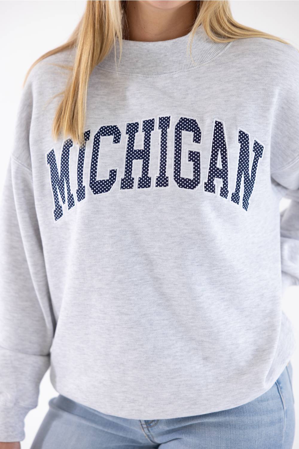 A close-up of a person wearing a light heather gray crewneck sweatshirt with 'MICHIGAN' printed across the chest in navy blue block letters with a white polka-dot pattern, paired with light blue jeans.