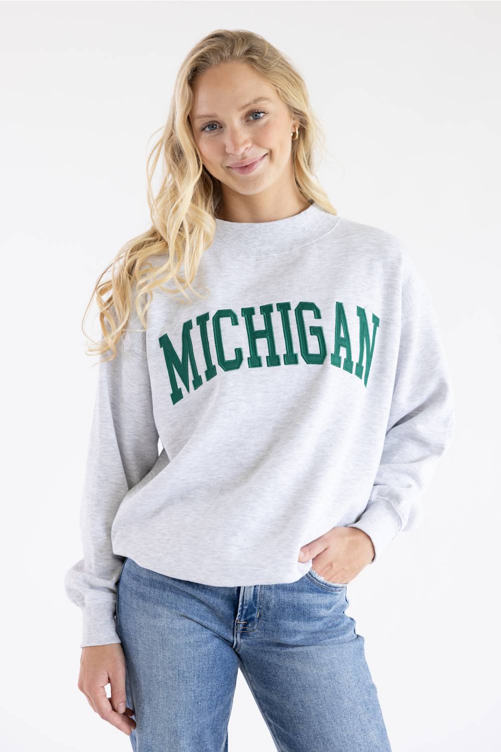 A woman modeling a light heather gray crewneck sweatshirt with 'MICHIGAN' printed in green varsity lettering across the chest, paired with blue jeans against a plain white background.