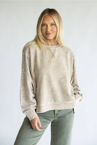 A woman modeling a beige acid-wash crewneck sweatshirt with unique sleeve cutouts, paired with olive green pants, against a plain light gray background.