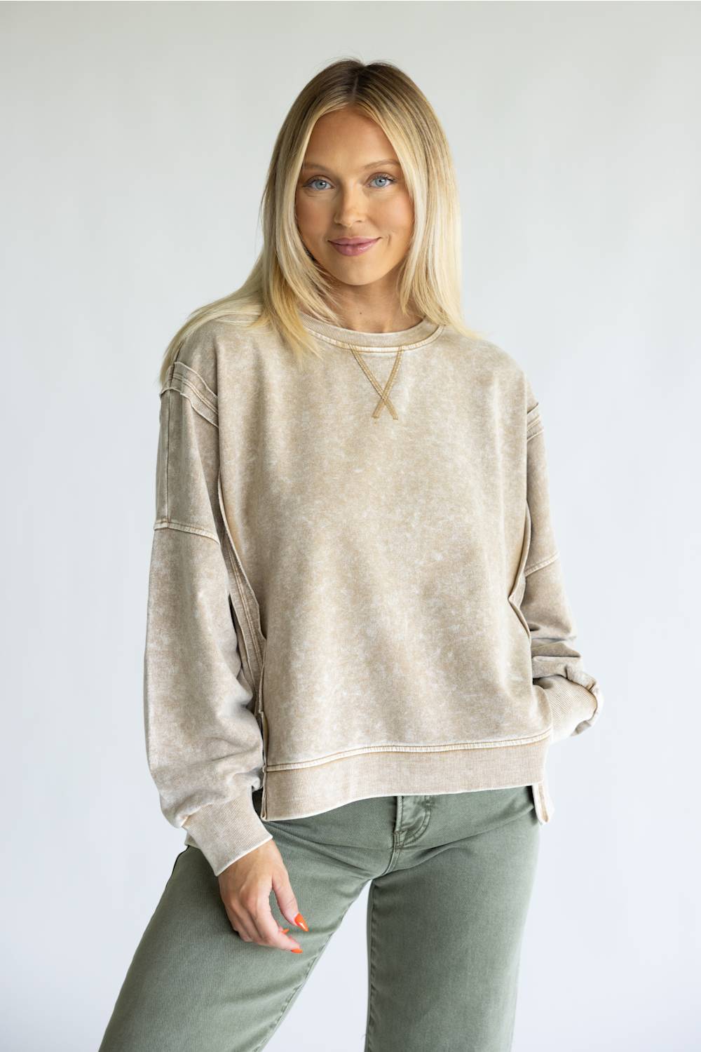 A woman modeling a beige acid-wash crewneck sweatshirt with unique sleeve cutouts, paired with olive green pants, against a plain light gray background.