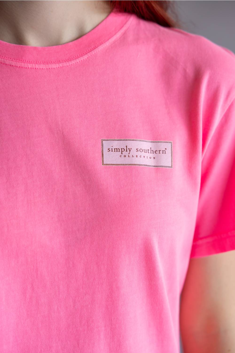A close-up of a person wearing a bright pink short-sleeve t-shirt with a small rectangular 'simply southern' collection logo patch on the left chest.