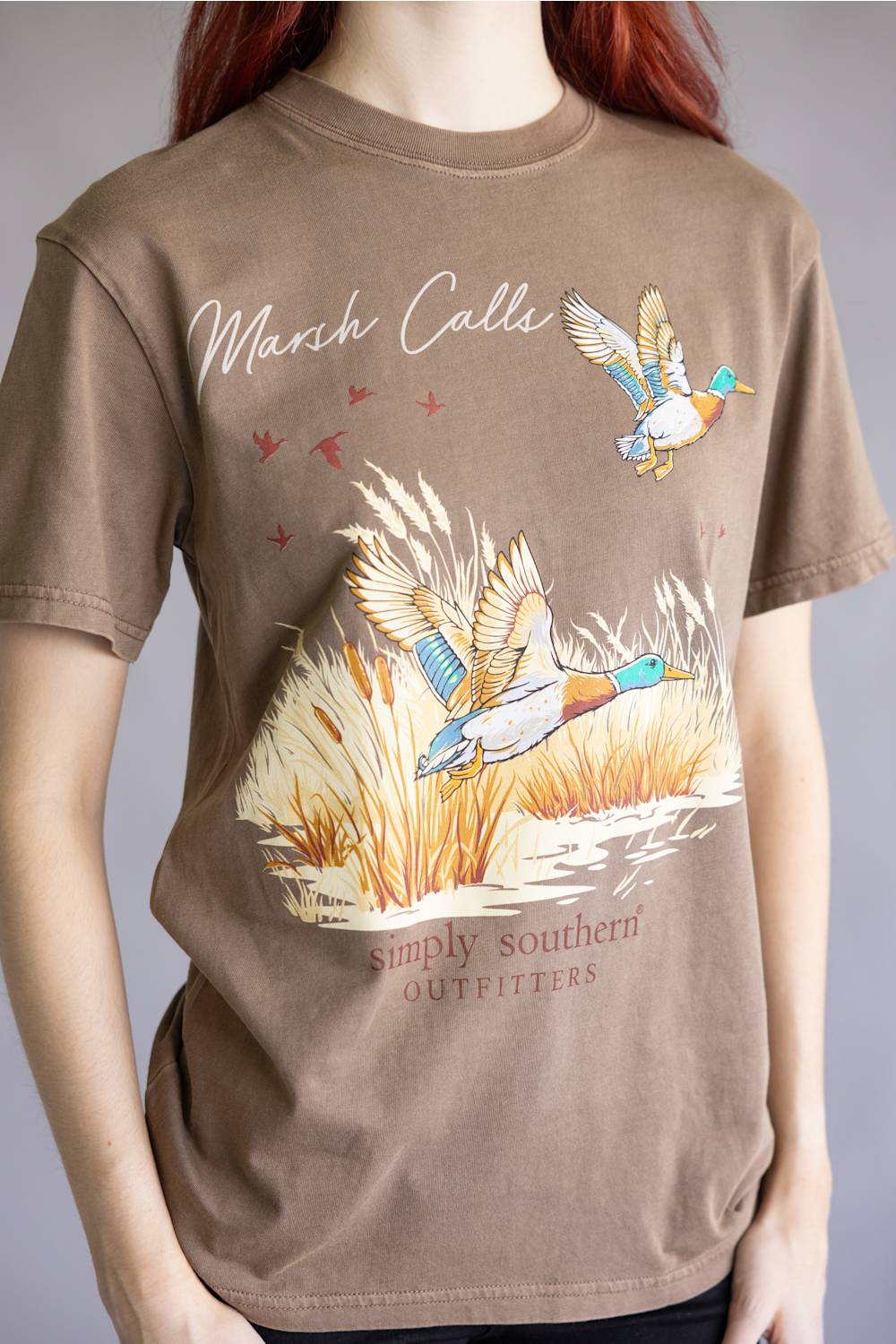 A person wearing a brown graphic t-shirt with a marsh duck illustration and the text 'Marsh Calls' and 'simply southern OUTFITTERS'.