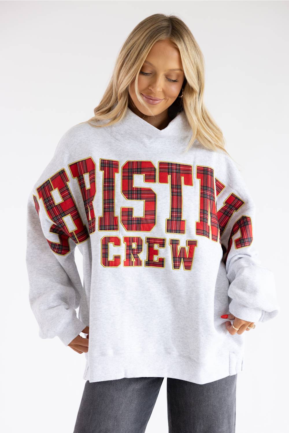 A woman wearing an oversized light gray sweatshirt with 'CHRISTINE CREW' printed across the front in red and black plaid lettering, paired with dark gray jeans, against a plain white studio background.