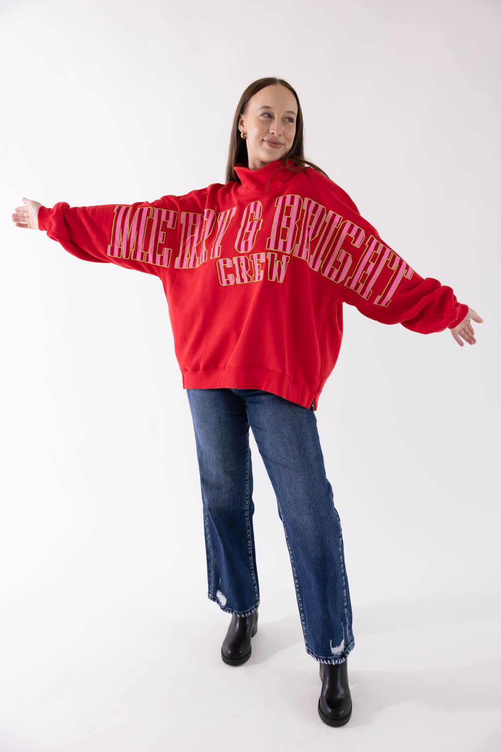 A woman modeling an oversized red hoodie with 'Merry & bright' printed in white across the front, paired with blue jeans and black boots, against a plain white studio background.