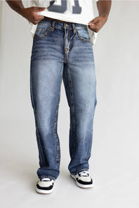 A person wearing loose-fit medium-wash blue denim jeans with a faded wash, paired with a white t-shirt and black-and-white sneakers, shown from the waist down against a light gray studio background.