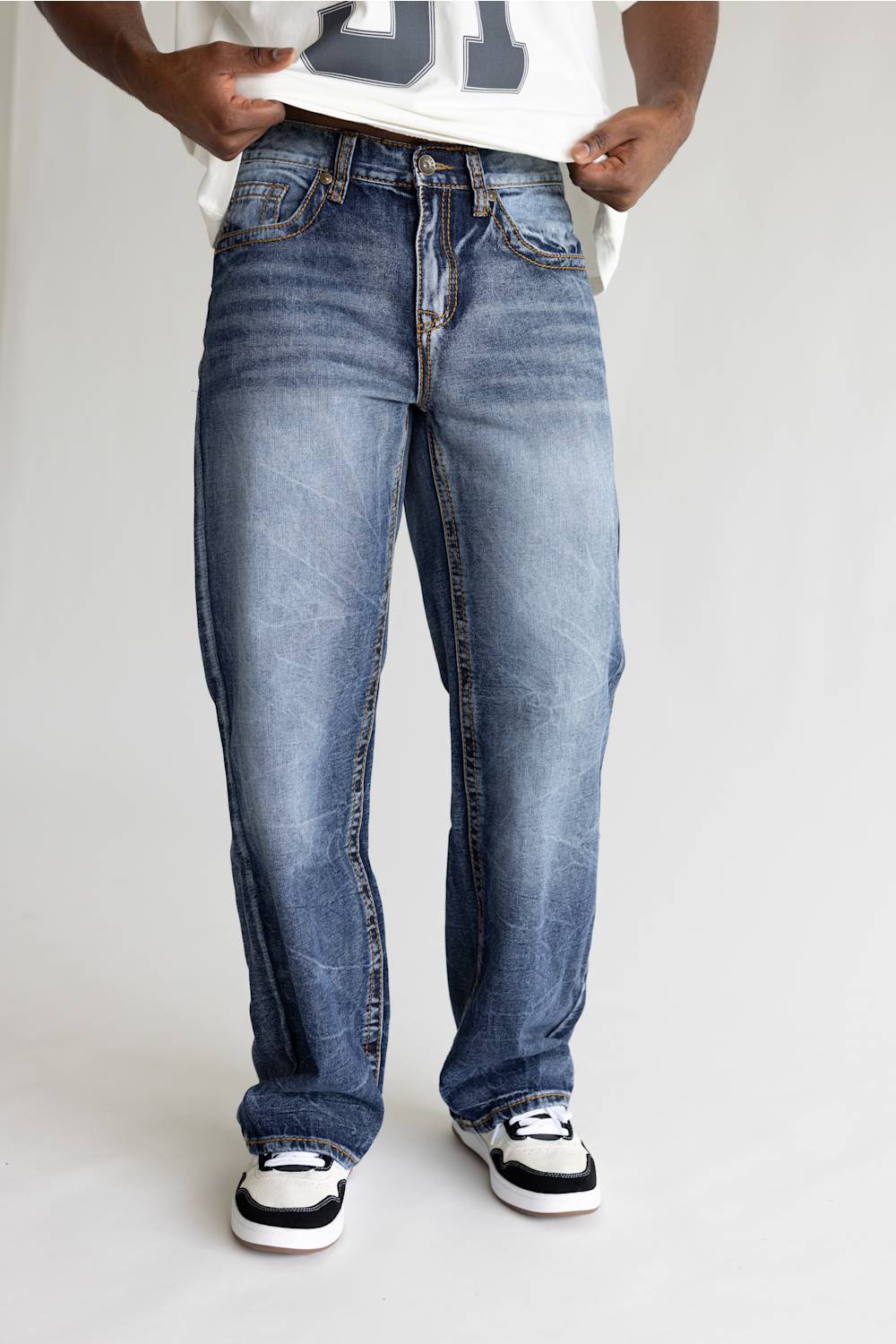A person wearing loose-fit medium-wash blue denim jeans with a faded wash, paired with a white t-shirt and black-and-white sneakers, shown from the waist down against a light gray studio background.