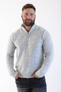 A man wearing a light gray marled knit quarter-zip sweater and dark jeans, standing against a plain white studio background.