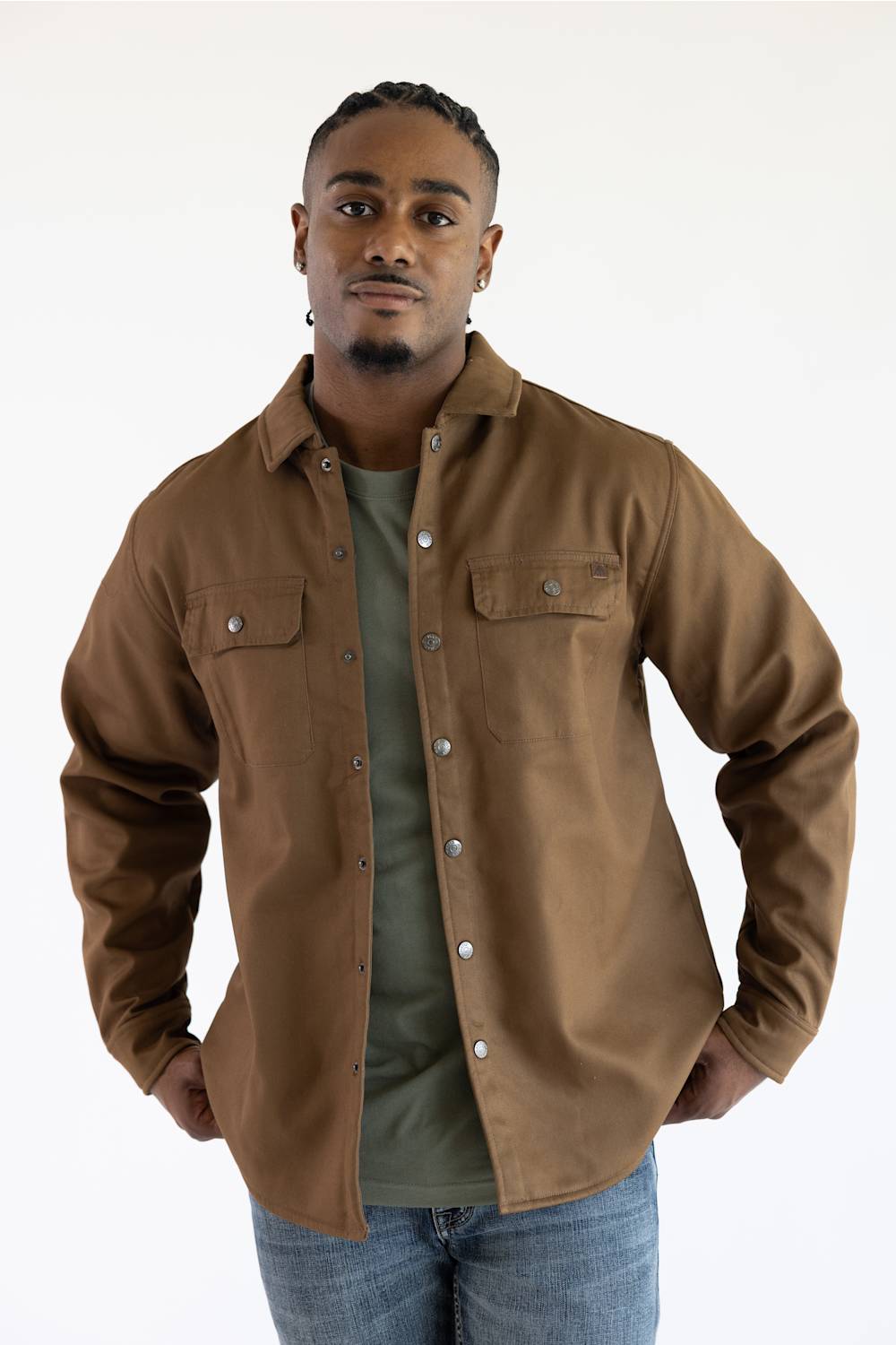A man wearing a brown button-up overshirt over an olive green t-shirt and blue jeans, standing against a plain white studio background.