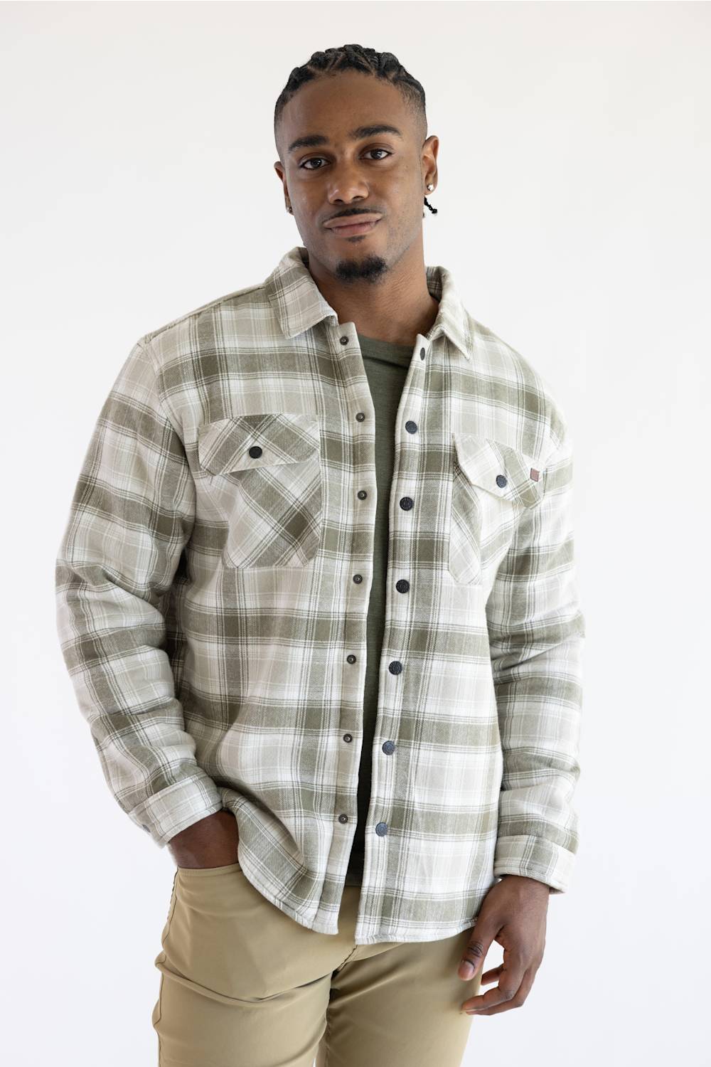A man wearing a long-sleeve plaid button-up shirt in beige and olive green, layered over a dark green t-shirt and khaki pants, standing against a plain white background.