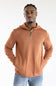 A man wearing a ribbed, quarter-zip pullover hoodie in a warm terracotta or rust orange color, paired with blue jeans, photographed against a clean white studio background.