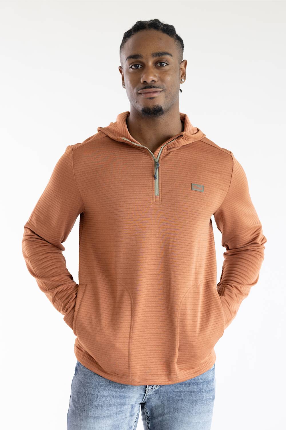 A man wearing a ribbed, quarter-zip pullover hoodie in a warm terracotta or rust orange color, paired with blue jeans, photographed against a clean white studio background.