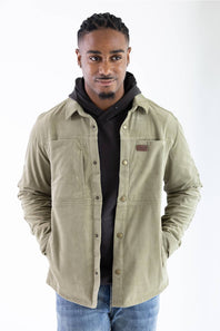 Front-facing upper-body portrait of a model wearing the Marsh Wear Delano Cord Shacket in Sage Brush green corduroy, featuring a dark hoodie underneath, antique brass snap buttons, a chest brand patch, and hands in the side pockets, styled with blue jeans.