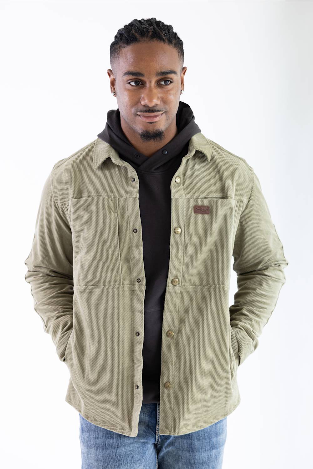Front-facing upper-body portrait of a model wearing the Marsh Wear Delano Cord Shacket in Sage Brush green corduroy, featuring a dark hoodie underneath, antique brass snap buttons, a chest brand patch, and hands in the side pockets, styled with blue jeans.
