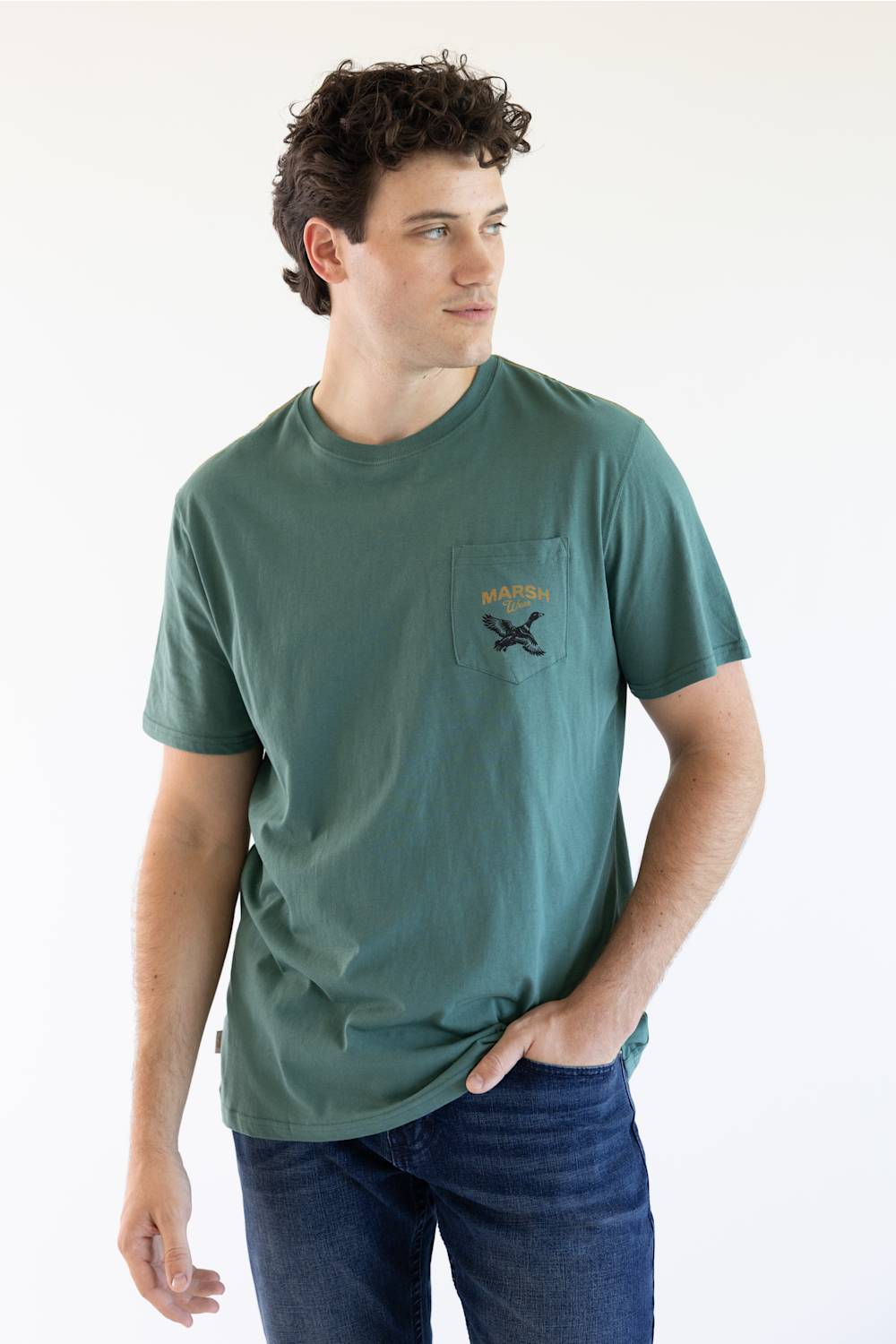 A man wearing a muted green pocket t-shirt with a small graphic on the chest pocket, paired with dark blue jeans, standing against a plain white background.