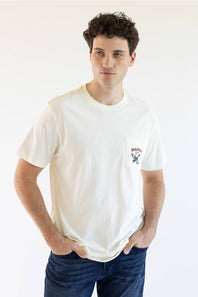 A man wearing a cream-colored short-sleeve pocket t-shirt with a small 'MARSHWEAR" graphic on the chest pocket, paired with dark blue jeans, against a plain white background.