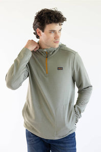 A man wearing a Marshwear hooded quarter-zip pullover in a textured sage green fabric with a small orange zipper and a small Marshwear  logo patch on the chest, paired with blue jeans against a plain white background.
