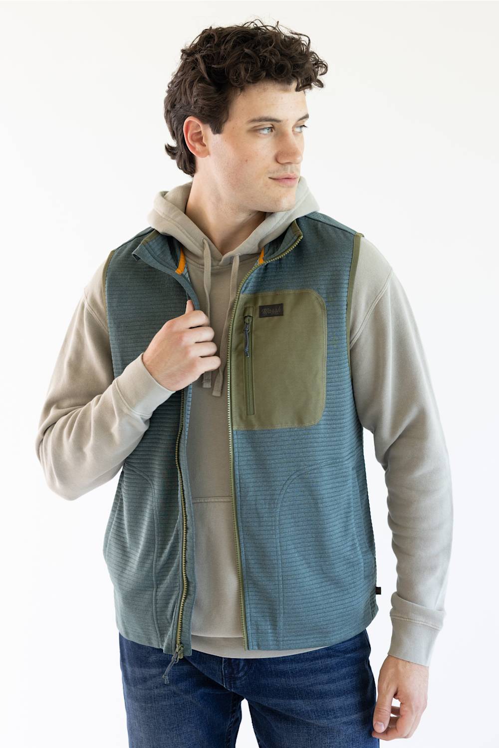 A man wearing a blue-green textured zip-up vest over a beige hoodie and blue jeans, photographed against a clean white studio background.