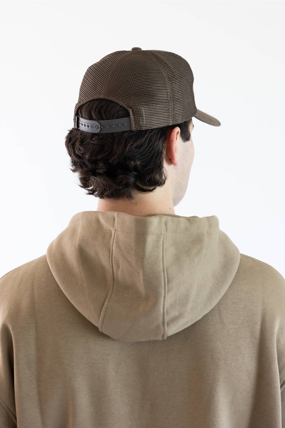 A man viewed from behind wearing a brown trucker-style cap and a beige hoodie against a plain white background.
