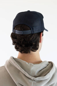 A man wearing a dark navy blue trucker hat with yellow embroidered 'Marsh Wear' text, paired with a light beige hoodie, against a plain white background.