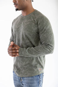Front studio portrait of a model wearing the long-sleeve Marsh Wear Surfside Waffle Knit Shirt in Fossil Foxhole Camo green, featuring a crew neck with a cross-stitch detail and waffle-textured fabric, styled with blue jeans.
