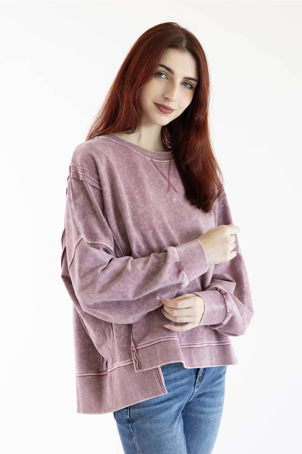 A woman with long red hair wearing a dusty rose acid-wash long-sleeve sweatshirt and blue jeans, standing against a plain white studio background.