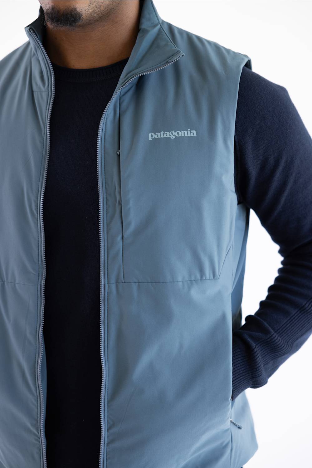A man wearing a Patagonia sleeveless zip-front vest in muted blue-gray over a dark navy long-sleeve shirt, shown from the lower face to the waist on a plain light background.