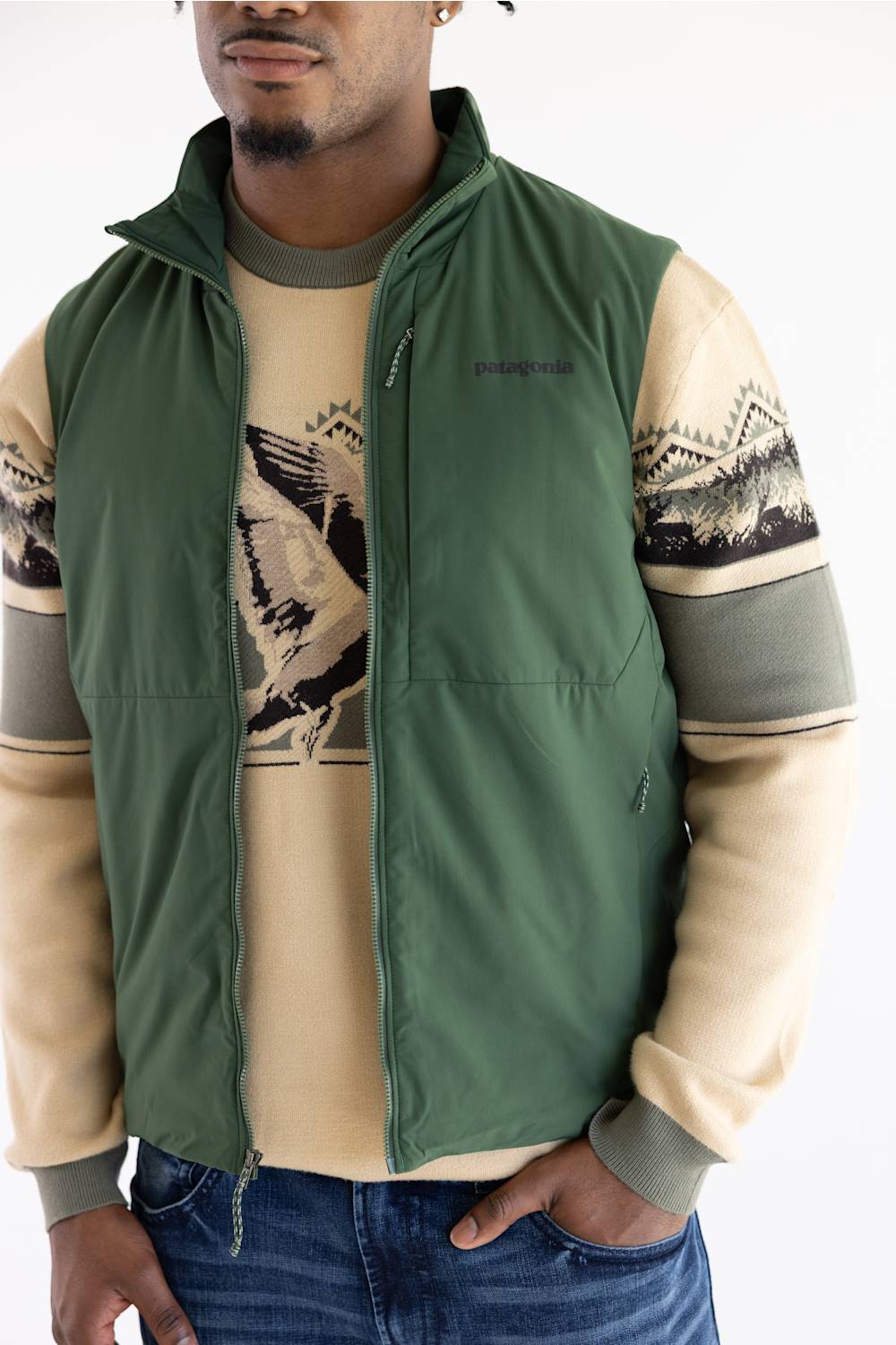 A man wearing a Patagonia zip-up vest over a beige and green patterned long-sleeve shirt, paired with blue jeans, against a plain white background.