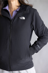 A close-up of a person wearing a black The North Face zip-front jacket over a light purple collared shirt, against a plain gray studio background.