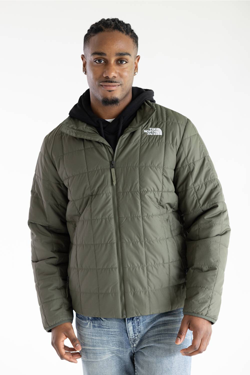 A man wearing an olive green quilted puffer jacket by The North Face, layered over a black hoodie and blue jeans, photographed against a plain white studio background.