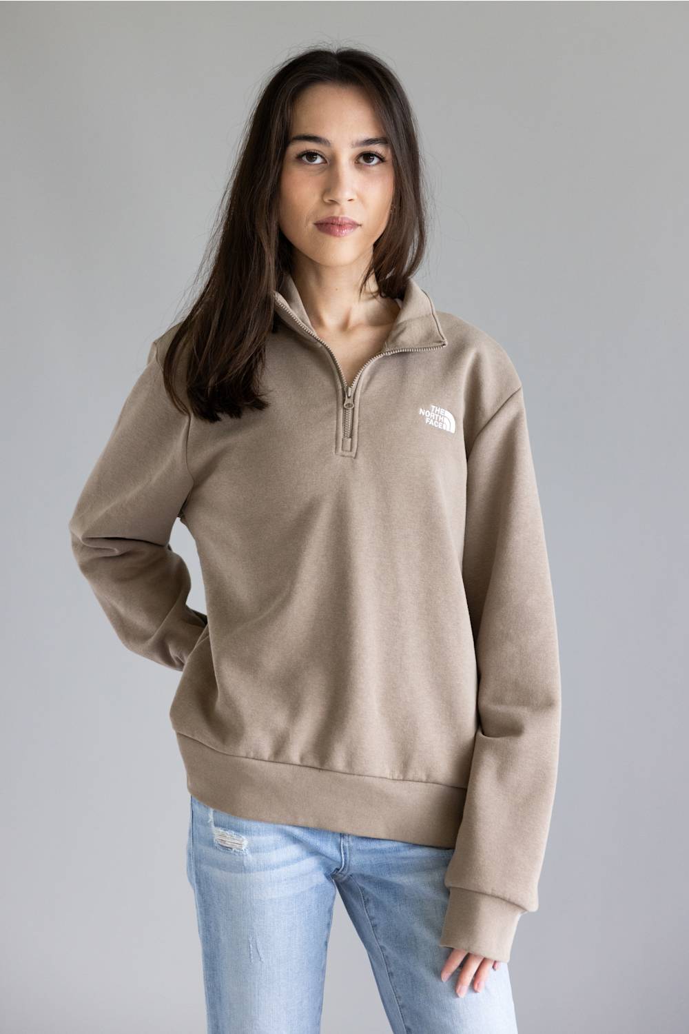 A woman wearing a taupe brown quarter-zip sweatshirt with a small white embroidered logo on the left chest, paired with light blue jeans, against a solid gray studio background.