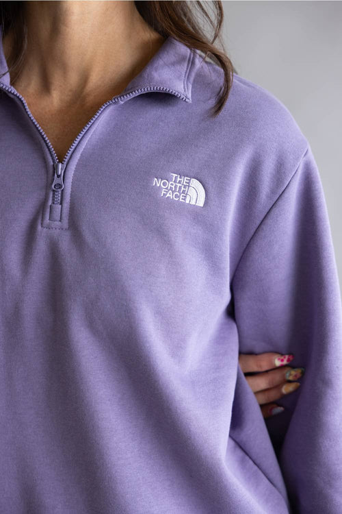 A close-up of a person wearing a lavender purple quarter-zip sweatshirt by The North Face, showing the white embroidered logo on the chest.