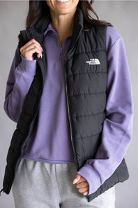A woman wearing a black quilted puffer vest by The North Face over a purple quarter-zip sweatshirt and gray sweatpants, shown against a plain gray studio background.