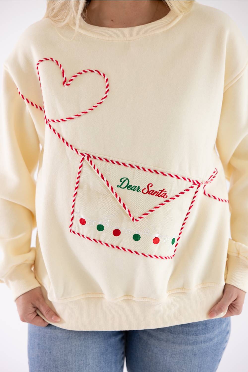 A cream-colored crewneck sweatshirt featuring a red and white rope-style graphic of an envelope with the words 'Dear Santa' in green script, worn by a model with blue jeans.