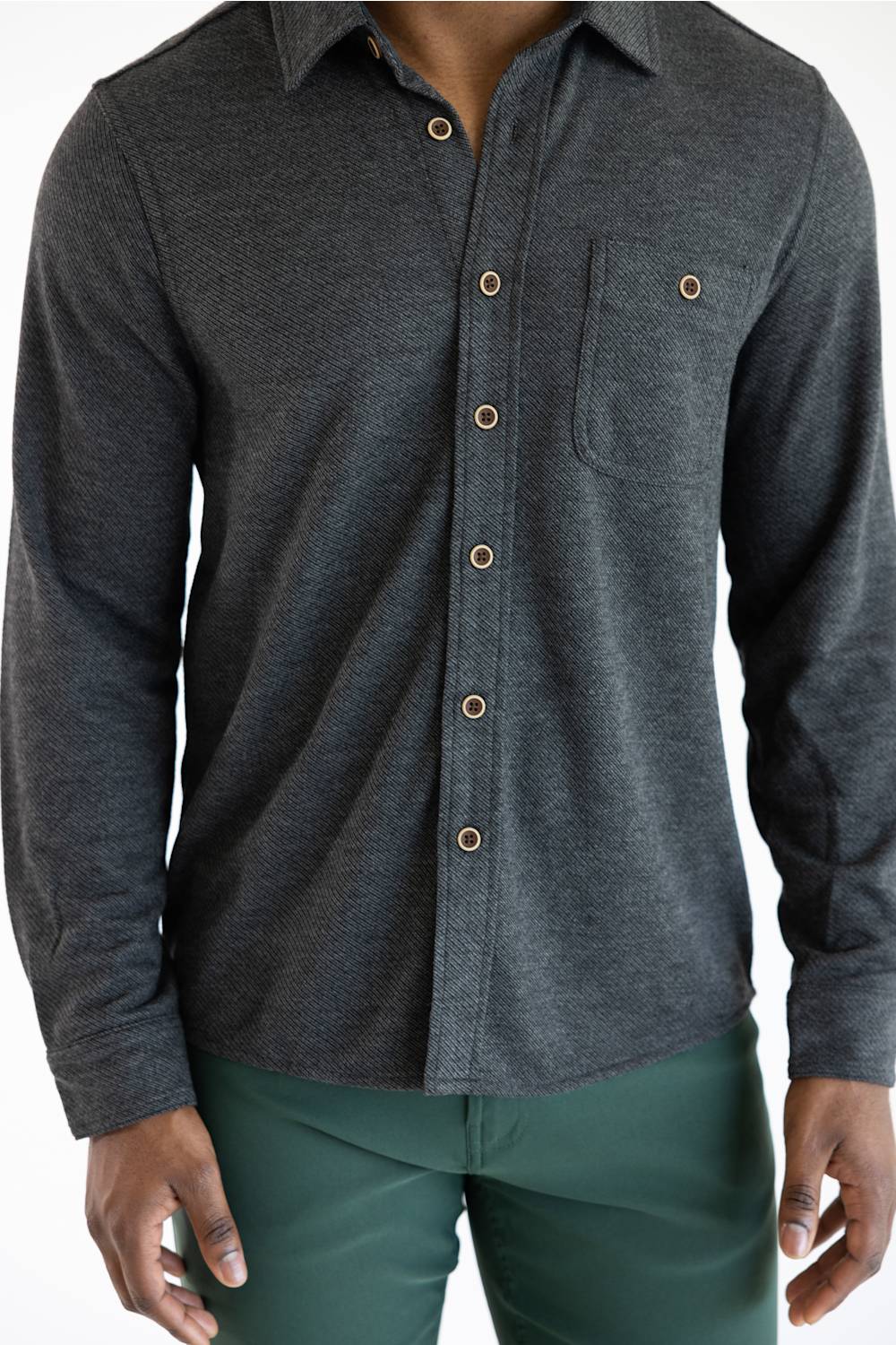 A man wearing a dark gray textured long-sleeve button-up shirt with brown buttons and a chest pocket, paired with green pants.