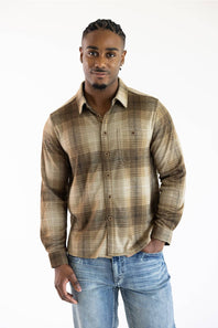 A man wearing a brown and beige plaid button-down shirt with blue jeans, standing against a plain white background.