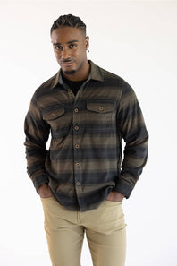 A man wearing a dark brown and black striped long-sleeve button-up flannel shirt with beige pants, standing against a plain white studio background.