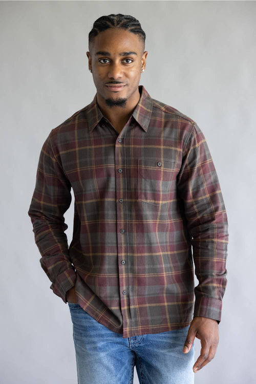 A man wearing a long-sleeve button-down plaid shirt in brown, red, and green tones, paired with blue jeans, standing against a neutral gray studio background.