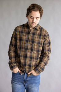 A man wearing a brown and black plaid button-down shirt with blue jeans, standing against a neutral gray studio background.