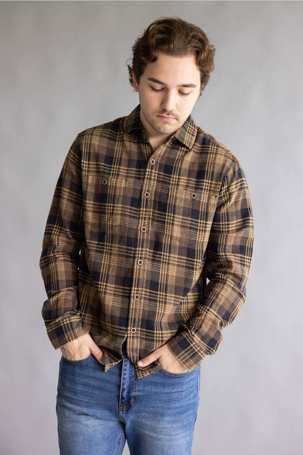A man wearing a brown and black plaid button-down shirt with blue jeans, standing against a neutral gray studio background.