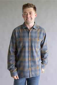 A smiling man wearing a long-sleeve plaid button-up shirt in blue and brown tones, paired with blue jeans, standing against a neutral gray studio background.